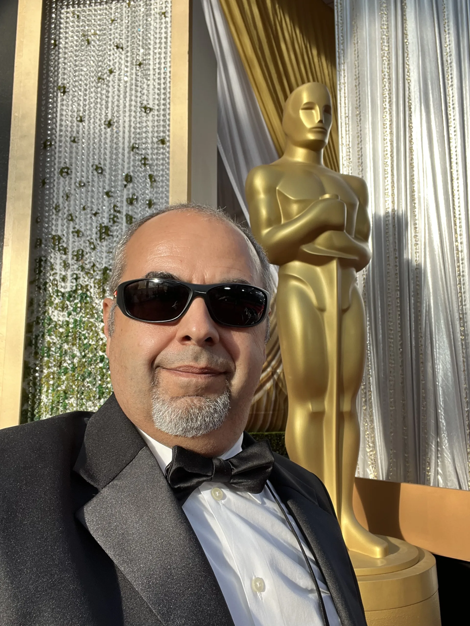 A man wearing sunglasses and a tuxedo in front of an oscar statue.